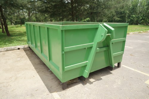 A large, rectangular green waste skip with reinforced side panels and a heavy-duty steel construction, situated on a paved surface in an outdoor area. The skip features a sturdy, angled door with a locking mechanism at the front, providing access for waste disposal. It is positioned on a wooden pallet base to allow for easier handling and drainage. The background includes a grassy area with dense trees and natural greenery, indicating an environment suitable for waste collection. The lighting suggests a clear day with natural sunlight, casting soft shadows around the skip. This image showcases a typical type of waste container used by commercial waste removal services, such as Commercial Waste Spitalfields, for collecting and transporting rubbish, with a focus on durability and practical urban placement in a park or residential setting.