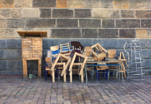 A collection of various wooden furniture pieces, including chairs, small tables, and stools, arranged against a brick wall on a paved outdoor surface. The furniture is in different shades of light and dark wood, with some appearing unfinished and others more polished. On the left side, there is a tall wooden cabinet or storage box with a slightly open lid, made of rough, light-colored wood. To the right of the furniture, a metal ladder is leaning against the wall, and the entire scene is set outdoors on an uneven cobblestone pavement, typical of a back alley or service area. The environment suggests a space used for temporary storage or collection of discarded furniture awaiting removal, aligning with commercial waste services in the Spitalfields area near London.