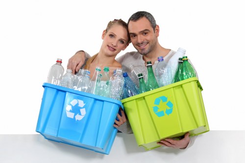 A smiling young couple stands close together, each holding a large plastic recycling bin; the woman's bin is blue and the man's is green, both with white recycling symbols on the front. The blue bin contains empty clear plastic bottles, some with caps on, and the green bin holds more bottles, mostly green and transparent, with a few white caps visible. They are positioned in front of a plain white background, suggesting an indoor setting, and are dressed casually, indicating a focus on recycling or waste management actions. The image highlights environmentally conscious waste segregation and recycling practices, fitting for a rubbish removal service in Spitalfields or nearby London areas. Commercial Waste Spitalfields promotes responsible waste handling, aligned with local recycling initiatives, with a visual emphasis on plastic bottle recycling. The scene's clarity, bright lighting, and focus on the recycling bins illustrate proper rubbish sorting, pertinent to professional waste removal services aimed at reducing environmental impact in urban environments like Spitalfields.