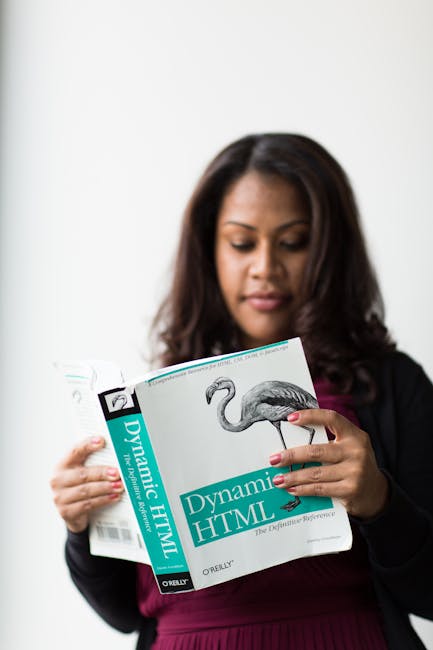 A woman with medium skin tone and long, dark brown hair wearing a black blazer and a dark pink top is standing indoors against a plain white background. She is holding an open book titled 'Dynamic HTM