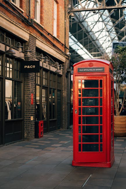 Centered in an indoor market setting, a promotional display features a vintage red telephone booth repurposed as a decorative stand for pumpkins and potted plants. The pumpkins, varying in size and sh